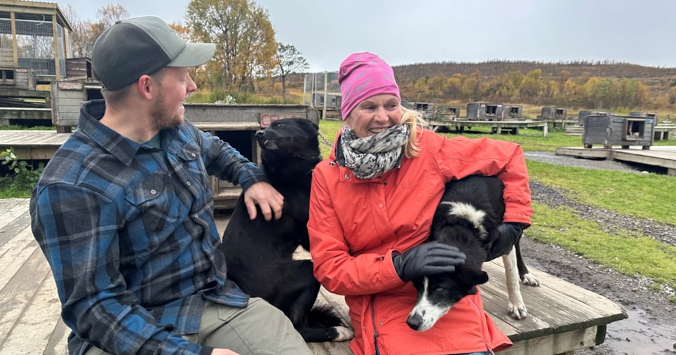 Kvinne og mann koser med to hunder foran en hundegård.