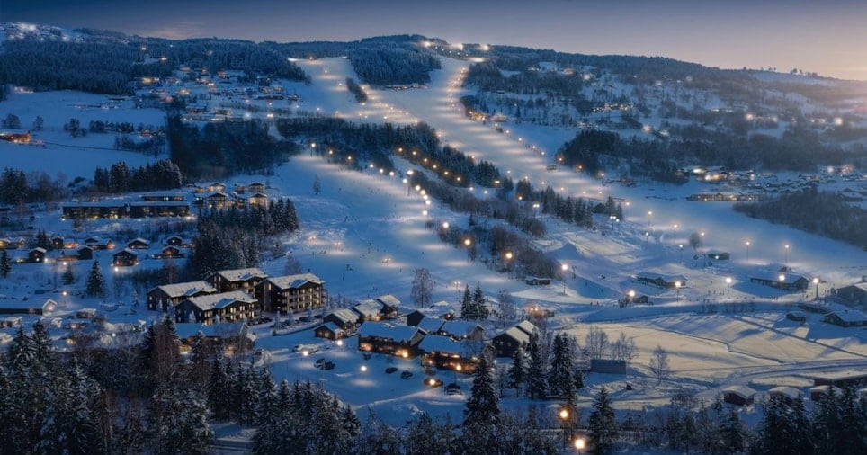 Et snødekket bygdelanskap i mørket, med lys i skibakken og hus, omringet av vakkert skoglandskap. Foto