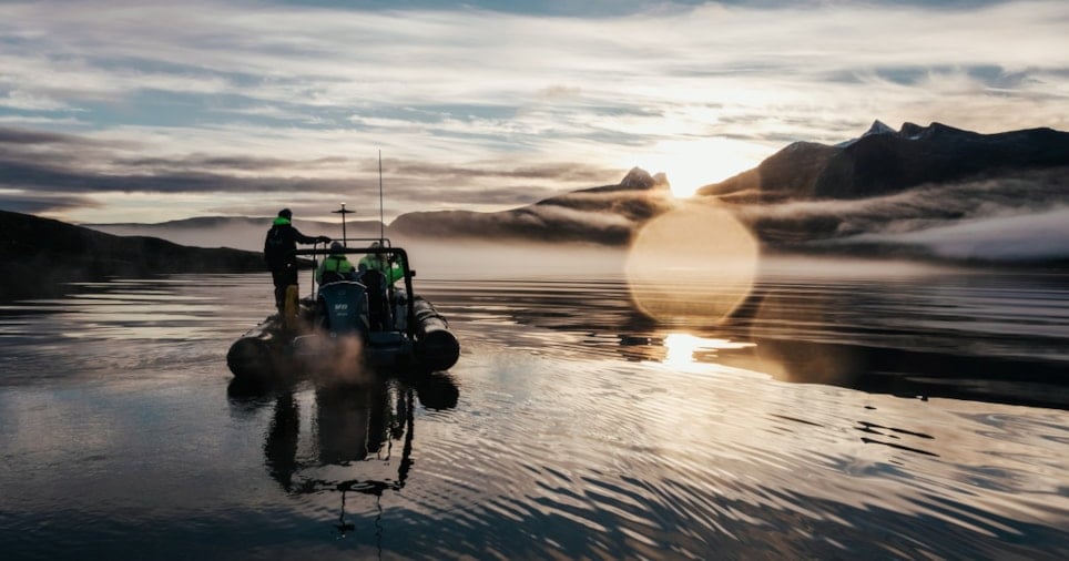 En gummibåt med tre personer ombord, i en stille fjord med tåke og fjell i soloppgang. Foto