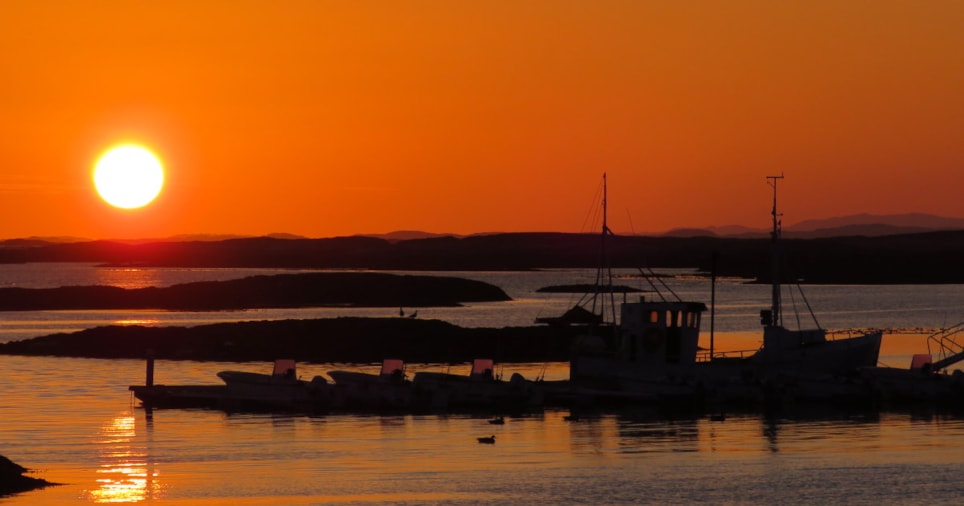 Silhuetter av båter og øyer mot en oransje himmel ved solnedgang. Foto.