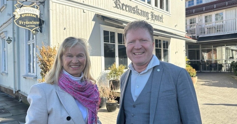 To smilende personer som står foran en hotellbygning i tre. På fasaden henger et skilt med Reenskaug Hotel på, og et skilt som viser til Reenskaug spisesal og en reklame lettøl. Foto