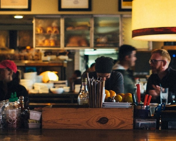 Bilde tatt fra bak en bardisk i en restaurant, med spisegjester i bakgrunnen. Foto.