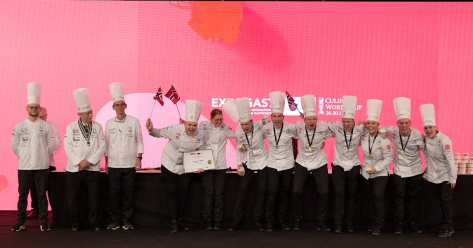 En rekke med veldig glade kokker i kokkeklær. Noen av dem holder norske flagg. Foto