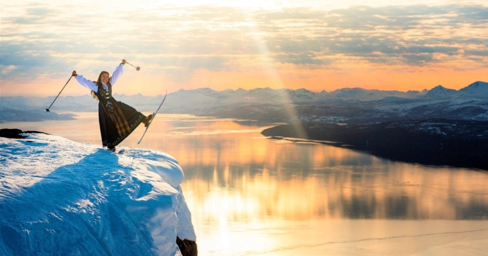 en glad jente på en topp av et fjell som har på seg ski og bunad