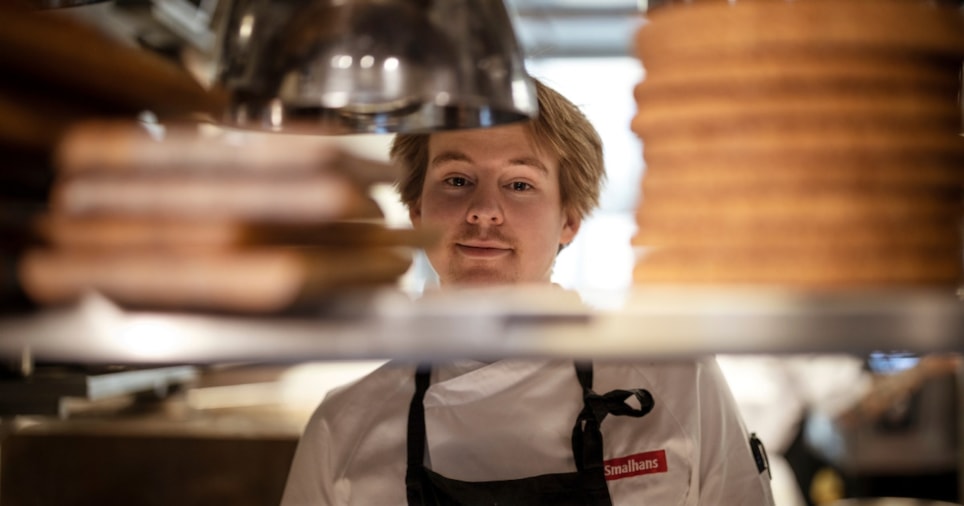 Ung kokk som titter mellom serveringshyller på et restaurantkjøkken. Foto