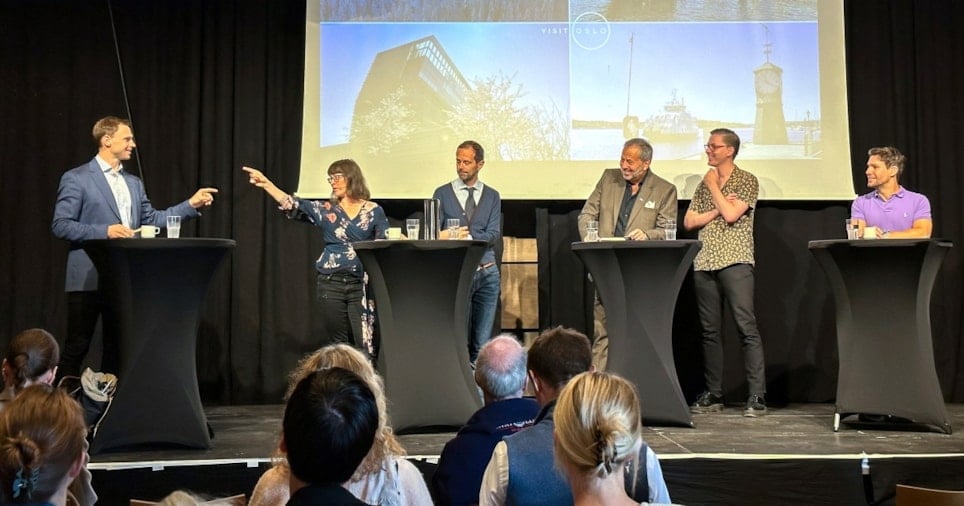 Seks smilende mennesker som står på en rekke på en scene med en presentasjon i bakgrunnen. Mannen og kvinnen som står ytterst til venstre i rekken peker på hverandre.